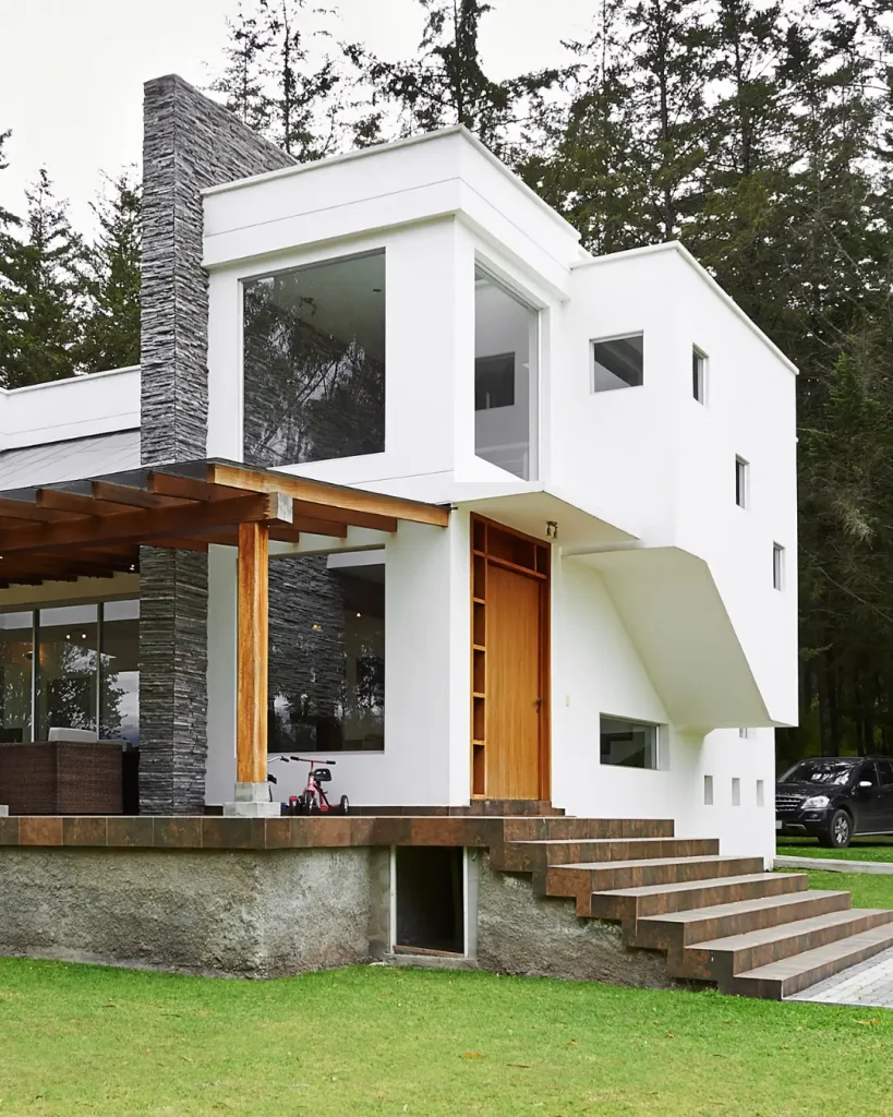 Contemporary two-story house with white walls, large glass windows, stone accents, and wooden finishes surrounded by greenery.