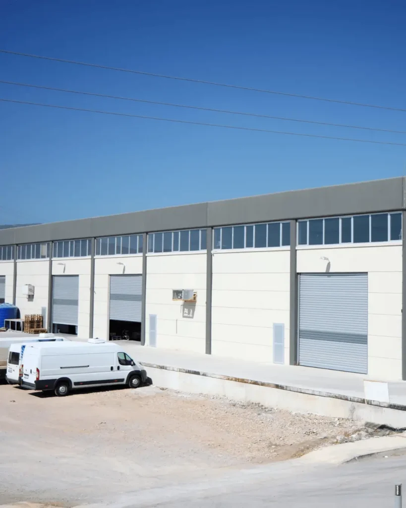 Modern industrial warehouse facility with roller doors and service vehicles parked outside.