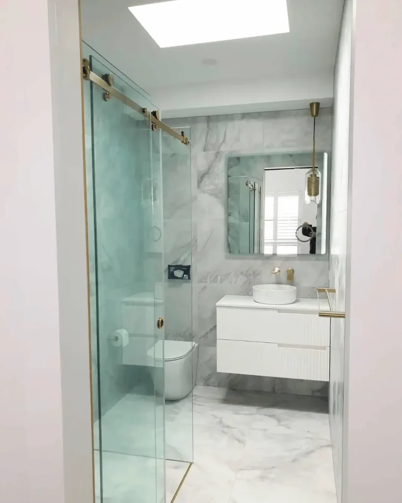 Sleek marble-clad bathroom with white fluted vanity, brass fittings, and walk-in glass shower under skylight