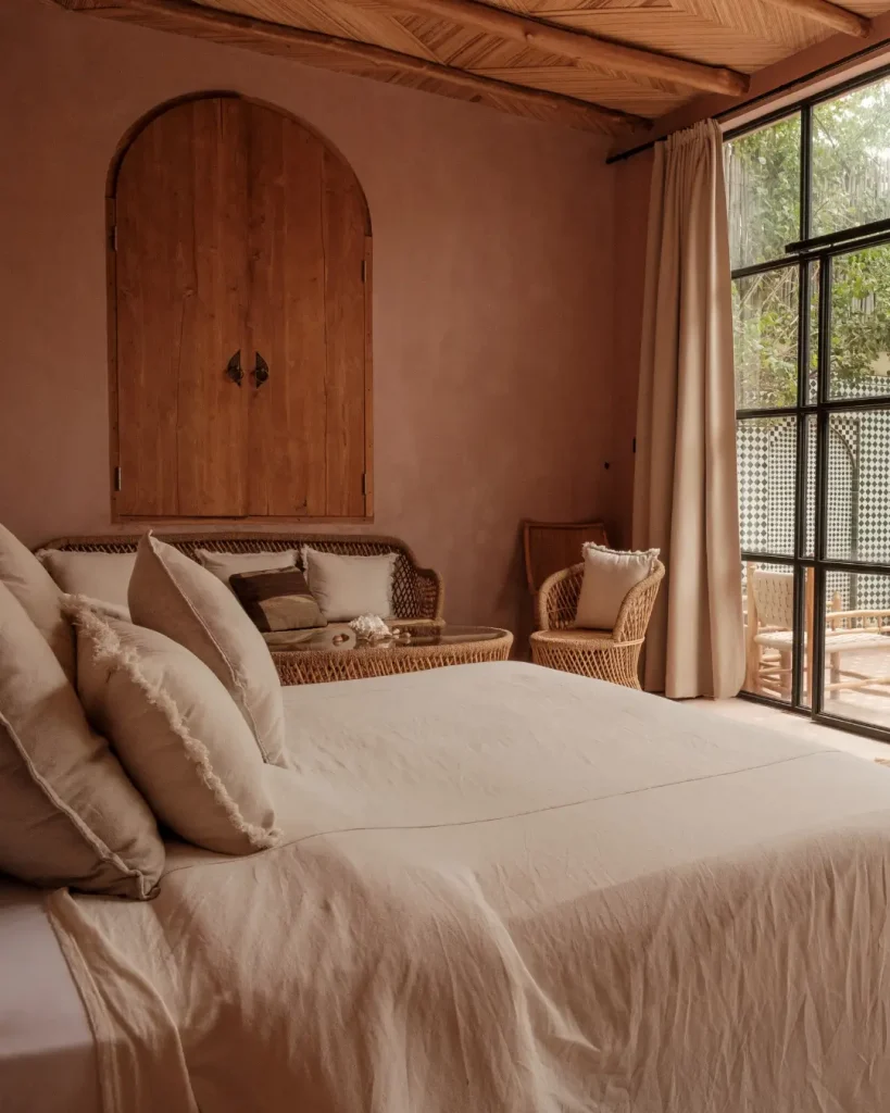Earth-toned outdoor bedroom addition with rattan furniture, soft linen bedding, and large glass doors opening to a patio.
