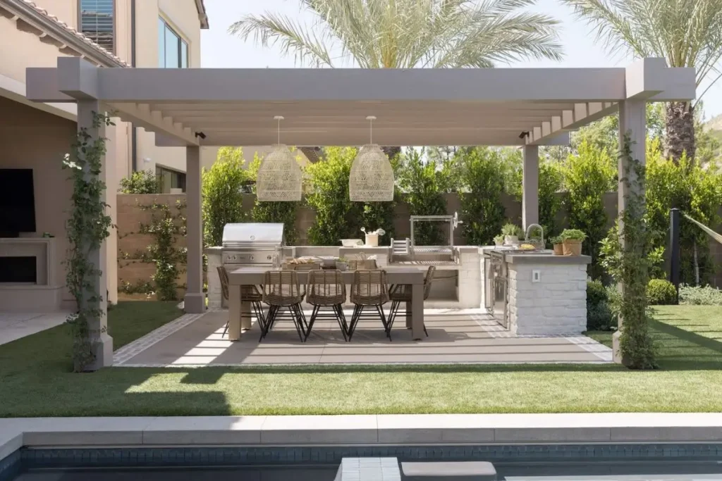 Modern outdoor kitchen and dining area with stone island, pergola roof, and wicker dining chairs overlooking a swimming pool.