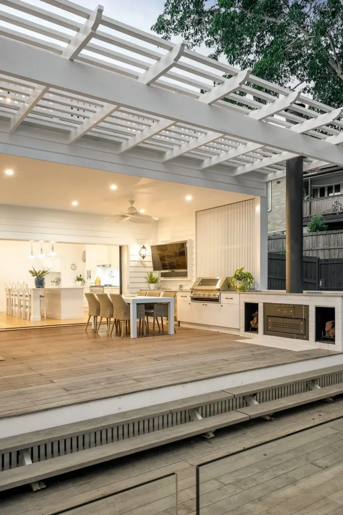 Luxury outdoor dining and kitchen area with timber deck, built-in BBQ, fireplace, and a white pergola roof.