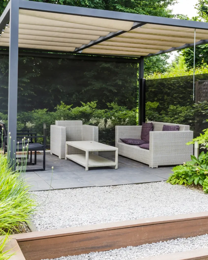 Outdoor lounge area with light wicker armchairs and coffee table under a modern pergola, surrounded by lush greenery and gravel landscaping.