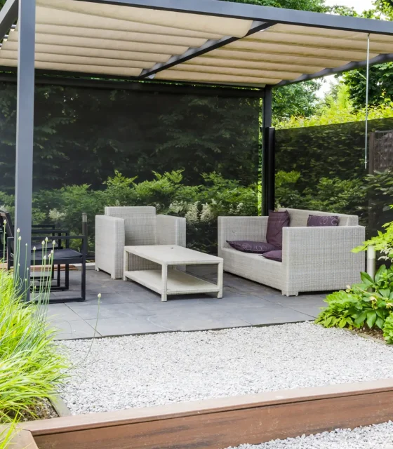 Outdoor lounge area with light wicker armchairs and coffee table under a modern pergola, surrounded by lush greenery and gravel landscaping.