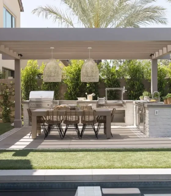Modern outdoor kitchen and dining area with stone island, pergola roof, and wicker dining chairs overlooking a swimming pool.